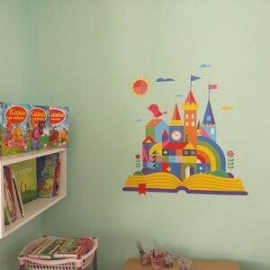 May include: A colorful illustration of a castle built on an open book with a rainbow arch, a sun, and a bird. The wall is painted a light green color. There is a white shelf with books on it.
