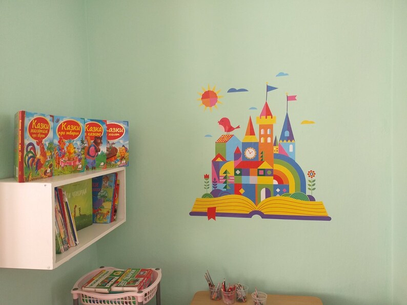 May include: A colorful illustration of a castle built on an open book with a rainbow arch, a sun, and a bird. The wall is painted a light green color. There is a white shelf with books on it.