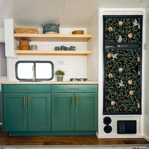 May include: A kitchen with green cabinets and a black refrigerator with a floral print. The refrigerator has a black door with a floral pattern and a black handle. The cabinets have gold handles. The kitchen has a white countertop and a white backsplash.