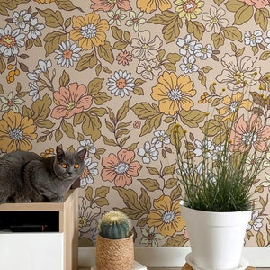 May include: A gray cat sits on a wooden shelf in front of a wall covered in a floral wallpaper with a brown background and yellow, orange, and white flowers. A cactus in a woven pot and a potted plant with green leaves are on the floor in front of the wall.