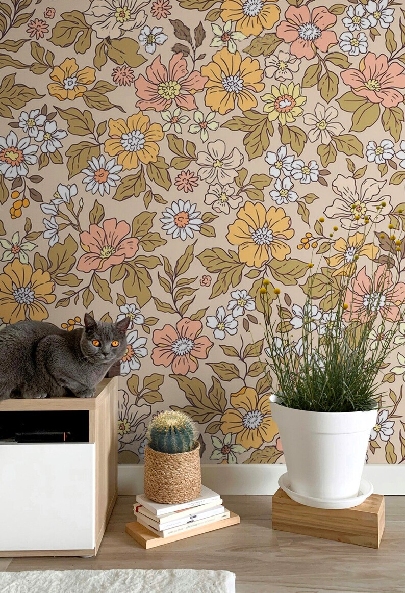May include: A gray cat sits on a wooden shelf in front of a wall covered in a floral wallpaper with a brown background and yellow, orange, and white flowers. A cactus in a woven pot and a potted plant with green leaves are on the floor in front of the wall.