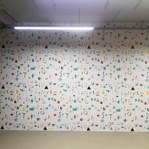 May include: A white wall with a repeating pattern of colorful letters, symbols, and illustrations. The letters are in a variety of fonts and sizes, and the symbols and illustrations are abstract and whimsical.