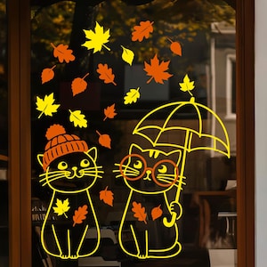 Könnte beinhalten: Ein Fensteraufkleber mit zwei Katzen in gelben Umrissen. Eine Katze trägt eine Strickmütze, die andere hält einen Regenschirm und trägt eine runde Brille. Das Design enthält verstreute gelbe und orangefarbene Herbstblätter.