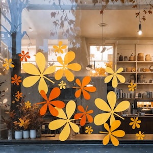 May include: A collection of yellow and orange leaf-shaped window decals applied to a glass surface. The decals feature various sizes of five-petaled flower or leaf silhouettes, creating a decorative autumn-themed display on the transparent pane.