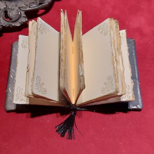 May include: An open miniature book with aged, cream-colored pages and decorative gold filigree. The book has a dark blue cover and a black tassel. The pages are slightly yellowed, giving it an antique appearance.