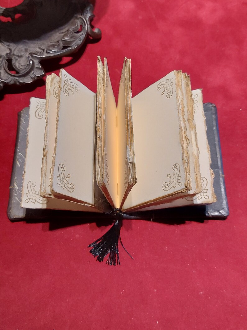 May include: An open miniature book with aged, cream-colored pages and decorative gold filigree. The book has a dark blue cover and a black tassel. The pages are slightly yellowed, giving it an antique appearance.