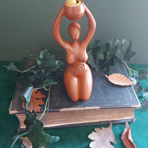 May include: A terracotta-colored incense holder in the shape of a stylized female figure, holding a small bowl aloft. The bowl contains a yellow incense cone. The figure is set on a vintage book, with decorative leaves around it.