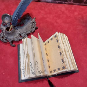 May include: An open, handmade journal with star and crescent moon designs on the pages. A black tassel bookmark hangs from the book. A blue feather quill pen rests in an ornate inkwell.