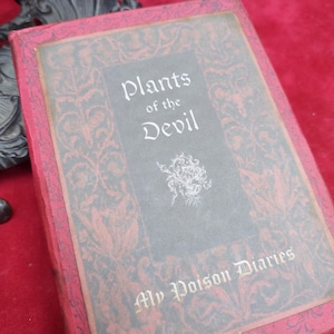 May include: A rectangular book with a red cover featuring a black ornate pattern and a central grey panel. The text reads Plants of the Devil and My Poison Diaries in a white serif font. A small white floral graphic sits below the main title.