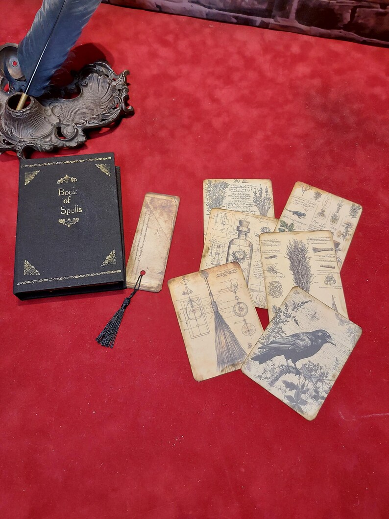 May include: A collection of fantasy-themed items on a red surface. Includes a black book titled "Book of Spells", a bookmark with a black tassel, and several aged-looking cards with botanical and crow illustrations. A quill and inkwell are in the background.