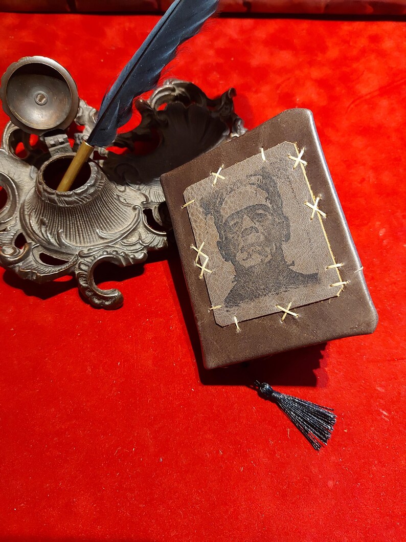 May include: A brown leather journal with a stitched-on image of Frankenstein's monster. The journal has a black tassel and is next to an ornate inkwell with a feather quill. The background is a solid red.