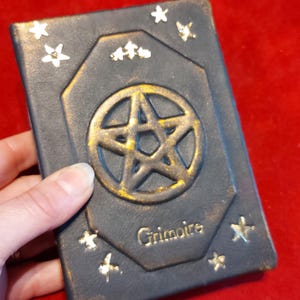 Puede incluir: Un grimorio azul oscuro con un pentagrama dorado y detalles de estrellas. La palabra "Grimoire" est&aacute; inscrita en letras doradas. El libro tiene un borde dorado y una borla negra. El libro est&aacute; sostenido por una mano.