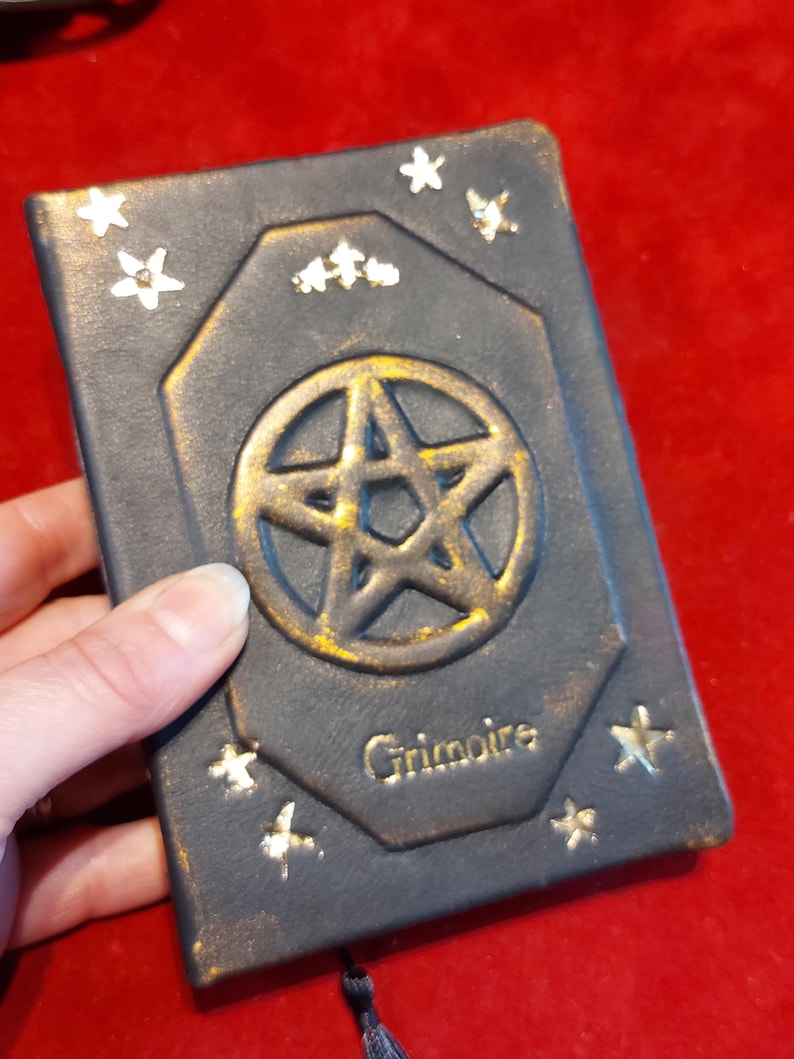 Puede incluir: Un grimorio azul oscuro con un pentagrama dorado y detalles de estrellas. La palabra "Grimoire" est&aacute; inscrita en letras doradas. El libro tiene un borde dorado y una borla negra. El libro est&aacute; sostenido por una mano.