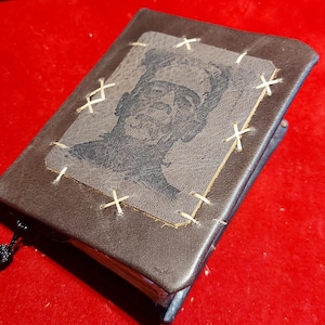 May include: A small, brown leather-bound journal with a stitched Frankenstein's monster image on the cover. The image is a black and white portrait of the monster. The journal has a black tassel attached to the spine.