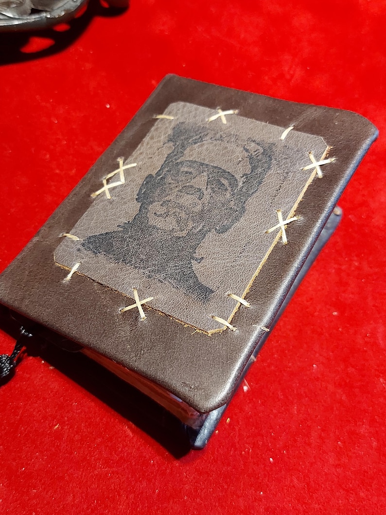 May include: A small, brown leather-bound journal with a stitched Frankenstein's monster image on the cover. The image is a black and white portrait of the monster. The journal has a black tassel attached to the spine.