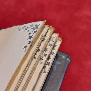 Puede incluir: Una pila de p&aacute;ginas de papel hechas a mano con bordes irregulares. Cada p&aacute;gina est&aacute; decorada con un borde de estrellas grises. Las p&aacute;ginas est&aacute;n encuadernadas con una cubierta gris oscuro, sobre un fondo rojo.