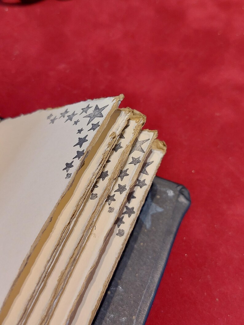 Puede incluir: Una pila de p&aacute;ginas de papel hechas a mano con bordes irregulares. Cada p&aacute;gina est&aacute; decorada con un borde de estrellas grises. Las p&aacute;ginas est&aacute;n encuadernadas con una cubierta gris oscuro, sobre un fondo rojo.