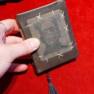 May include: A small, brown leather-bound book with a black tassel. The cover features a black and white image of Frankenstein's monster, stitched onto the cover with tan thread. The book is held against a red background.