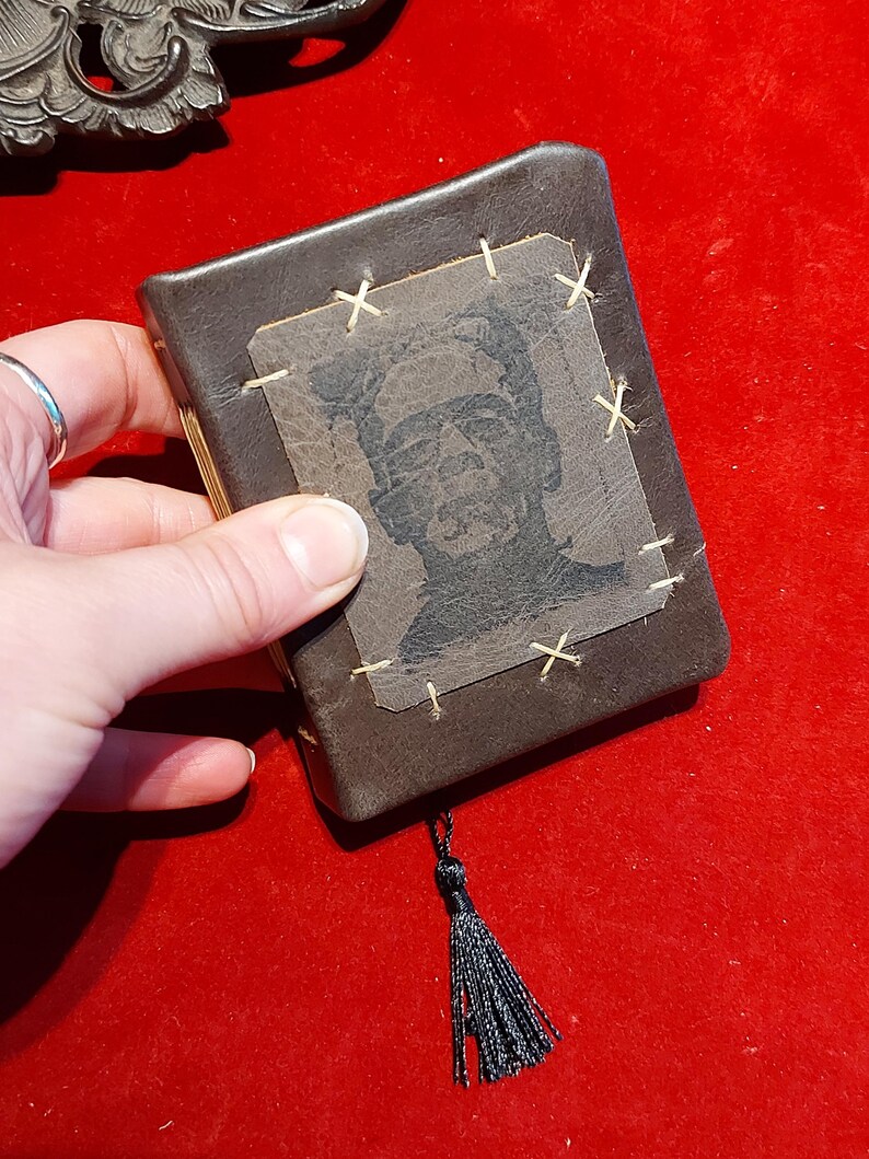 May include: A small, brown leather-bound book with a black tassel. The cover features a black and white image of Frankenstein's monster, stitched onto the cover with tan thread. The book is held against a red background.