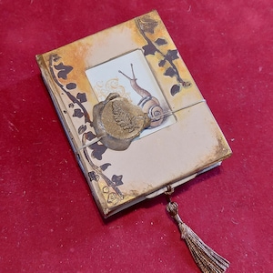 Puede incluir: Un diario hecho a mano con una ilustración de caracol en la portada, sellado con un sello de cera de bronce. El diario tiene una borla decorativa y presenta acentos botánicos en tonos marrón y dorado.
