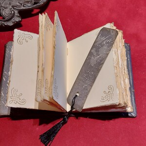 May include: An open, antique-style journal with aged, cream-colored pages and decorative corner flourishes. A metal bookmark with script hangs from the journal, and a black tassel is attached. The journal is on a red surface.