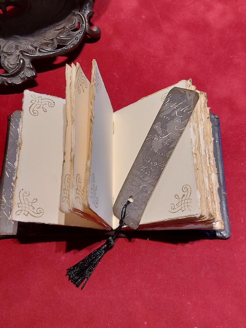 May include: An open, antique-style journal with aged, cream-colored pages and decorative corner flourishes. A metal bookmark with script hangs from the journal, and a black tassel is attached. The journal is on a red surface.
