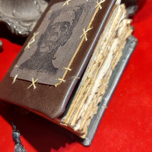 May include: A small, handmade leather-bound book with a portrait on the cover. The brown cover is stitched with light-colored thread. The pages are aged and rough-edged. A dark tassel hangs from the spine. The book is open, revealing the aged pages.