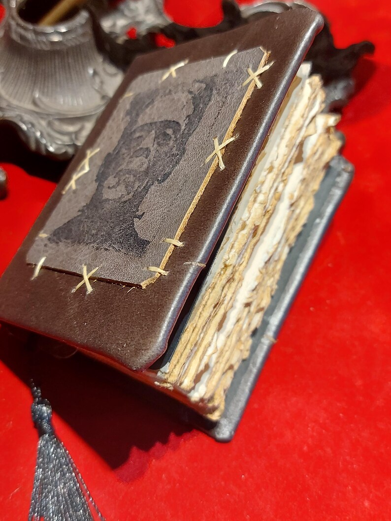 May include: A small, handmade leather-bound book with a portrait on the cover. The brown cover is stitched with light-colored thread. The pages are aged and rough-edged. A dark tassel hangs from the spine. The book is open, revealing the aged pages.