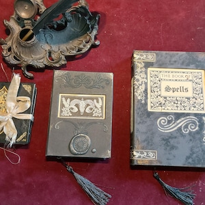 Puede incluir: Tres diarios negros con detalles dorados y cubiertas decorativas. Los diarios est&aacute;n titulados "El Libro de los Hechizos".