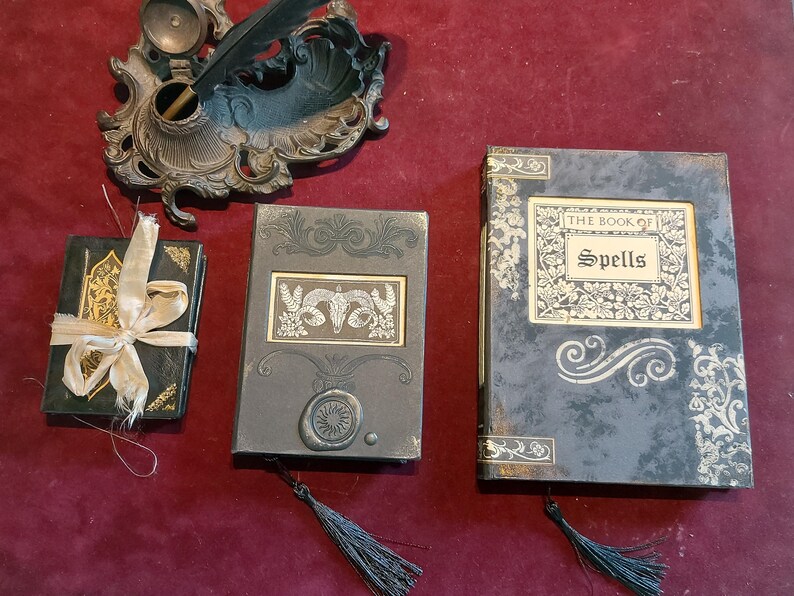 Puede incluir: Tres diarios negros con detalles dorados y cubiertas decorativas. Los diarios est&aacute;n titulados "El Libro de los Hechizos".