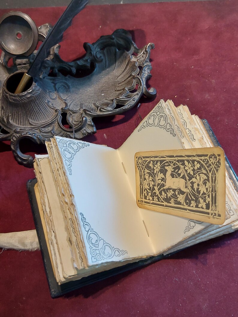 Puede incluir: Un diario antiguo abierto con una cubierta negra y p&aacute;ginas envejecidas. El diario est&aacute; decorado con dise&ntilde;os ornamentados y un marcap&aacute;ginas con una ilustraci&oacute;n en blanco y negro de un ciervo en un patr&oacute;n floral.