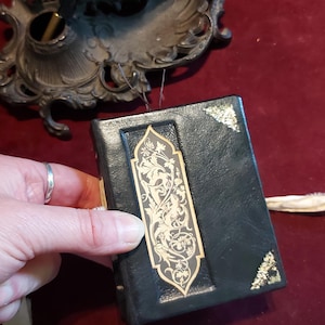 Puede incluir: Un peque&ntilde;o diario encuadernado en cuero negro con detalles dorados y un intrincado dise&ntilde;o floral en la portada.