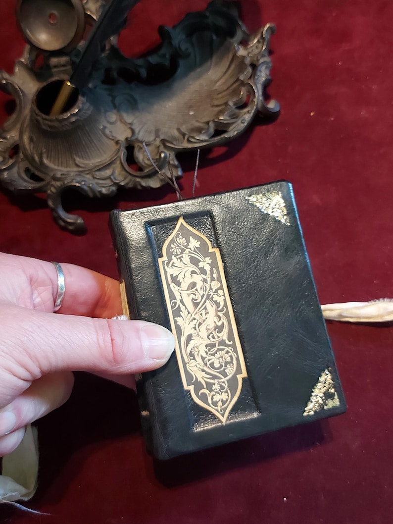 Puede incluir: Un peque&ntilde;o diario encuadernado en cuero negro con detalles dorados y un intrincado dise&ntilde;o floral en la portada.