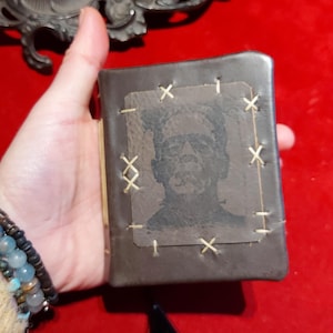 May include: A small, dark brown leather-bound book featuring a stitched Frankenstein's monster image on the cover. The cover is stitched with cream-colored thread. A black tassel hangs from the bottom of the book.