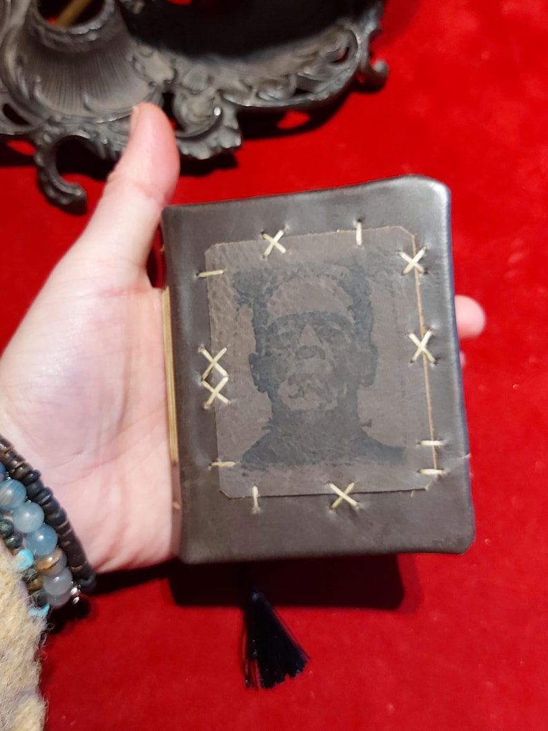 May include: A small, dark brown leather-bound book featuring a stitched Frankenstein's monster image on the cover. The cover is stitched with cream-colored thread. A black tassel hangs from the bottom of the book.
