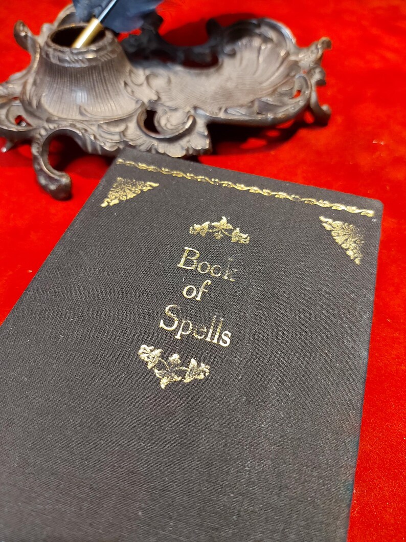 May include: A black book titled "Book of Spells" with gold lettering and decorative accents. An ornate, antique inkwell and quill are in the background. The objects are set against a red background.