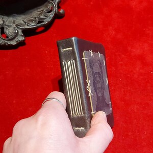 May include: A small, dark brown leather-bound journal with gold-colored stitching along the spine. The cover features a faded image of a face. The journal is held against a red background.