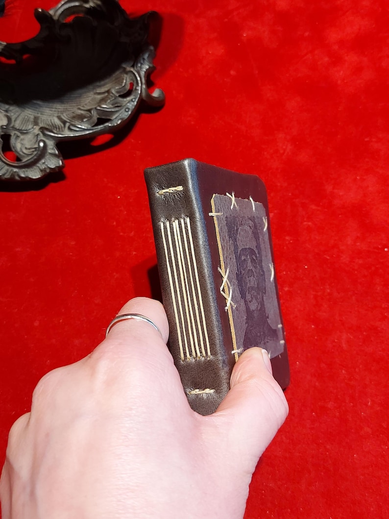 May include: A small, dark brown leather-bound journal with gold-colored stitching along the spine. The cover features a faded image of a face. The journal is held against a red background.