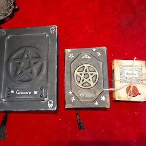Puede incluir: Tres libros decorativos con tem&aacute;tica oculta. El m&aacute;s grande es gris oscuro con un pentagrama y la palabra "Grimoire". El libro del medio es marr&oacute;n con un pentagrama dorado y el mismo texto. Tambi&eacute;n hay un peque&ntilde;o libro atado con hilo.