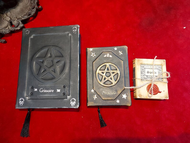 Puede incluir: Tres libros decorativos con tem&aacute;tica oculta. El m&aacute;s grande es gris oscuro con un pentagrama y la palabra "Grimoire". El libro del medio es marr&oacute;n con un pentagrama dorado y el mismo texto. Tambi&eacute;n hay un peque&ntilde;o libro atado con hilo.