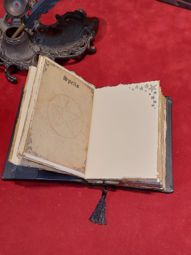 Puede incluir: Un libro abierto de estilo antiguo con la palabra "Spells" en la p&aacute;gina superior. Las p&aacute;ginas est&aacute;n envejecidas con ilustraciones de pentagramas y estrellas. Una borla negra cuelga del lomo. El libro est&aacute; sobre una superficie roja.