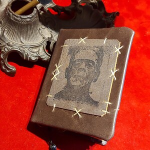 May include: A brown leather-bound journal with a stitched-on image of Frankenstein's monster. The image is stitched with gold thread. A black tassel hangs from the bottom. An antique inkwell and quill are in the background.