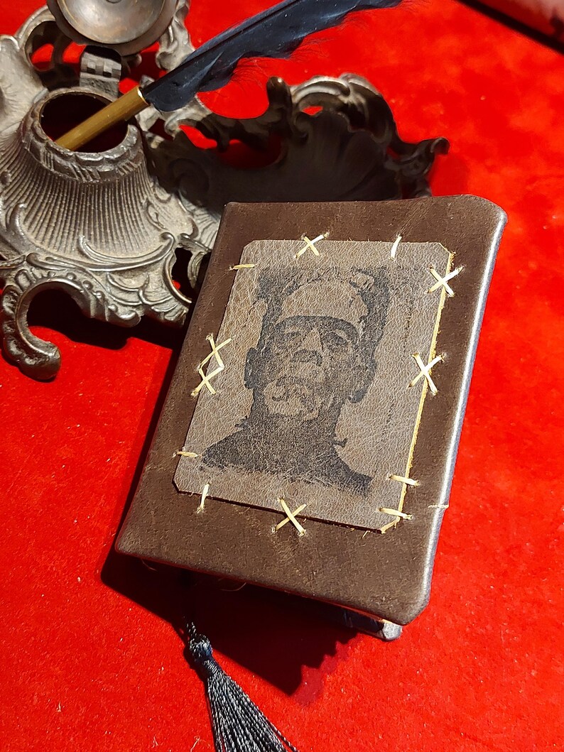 May include: A brown leather-bound journal with a stitched-on image of Frankenstein's monster. The image is stitched with gold thread. A black tassel hangs from the bottom. An antique inkwell and quill are in the background.