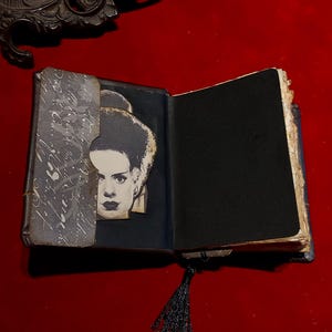 May include: An open, antique-style book with a black interior and a black tassel bookmark. A black and white image of a woman's face is visible on the left page. The book is on a red surface.