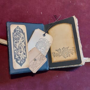 Puede incluir: Un peque&ntilde;o diario encuadernado en cuero negro con un dise&ntilde;o floral decorativo en la portada. El diario est&aacute; abierto para revelar una p&aacute;gina en blanco con un borde decorativo. Hay tres etiquetas en blanco con dise&ntilde;os florales adjuntas al diario.