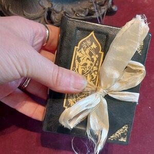 Puede incluir: Un peque&ntilde;o libro encuadernado en cuero negro con detalles dorados y un dise&ntilde;o floral decorativo. El libro est&aacute; atado con una cinta de color crema.