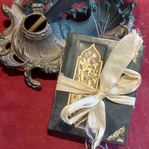 Puede incluir: Un peque&ntilde;o diario encuadernado en cuero negro con detalles dorados y una cinta de color crema atada alrededor. El diario est&aacute; sobre una superficie roja junto a un tintero antiguo y una pluma.