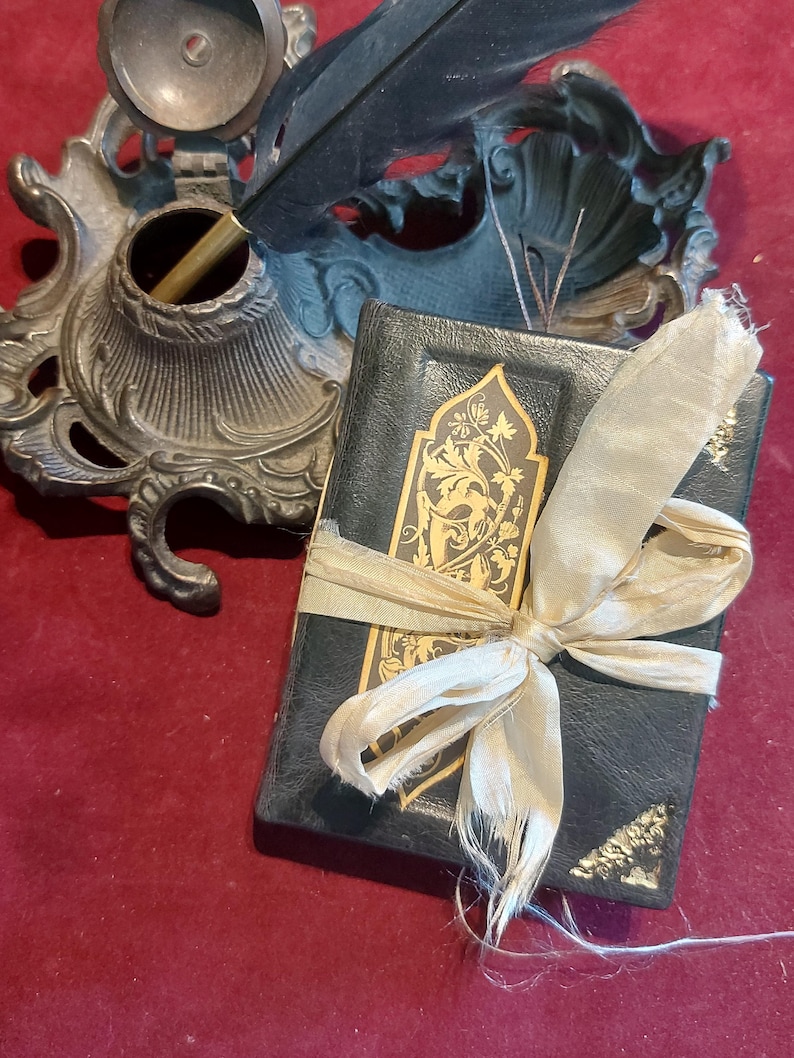 Puede incluir: Un peque&ntilde;o diario encuadernado en cuero negro con detalles dorados y una cinta de color crema atada alrededor. El diario est&aacute; sobre una superficie roja junto a un tintero antiguo y una pluma.