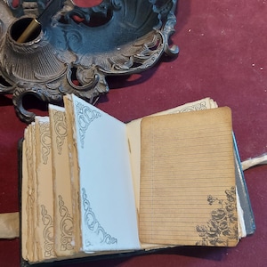 Puede incluir: Un peque&ntilde;o diario encuadernado en cuero negro con un cierre de metal decorativo. El diario est&aacute; abierto para revelar p&aacute;ginas rayadas con un dise&ntilde;o floral en la p&aacute;gina derecha. Una pluma y un tintero son visibles en el fondo.