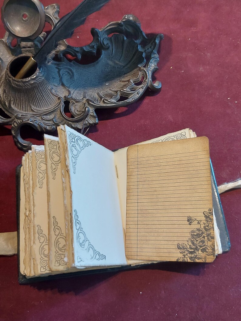 Puede incluir: Un peque&ntilde;o diario encuadernado en cuero negro con un cierre de metal decorativo. El diario est&aacute; abierto para revelar p&aacute;ginas rayadas con un dise&ntilde;o floral en la p&aacute;gina derecha. Una pluma y un tintero son visibles en el fondo.